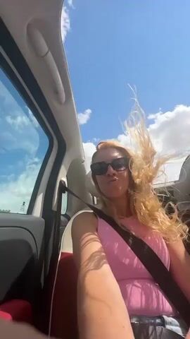 The video shows a woman with long blonde hair and sunglasses sitting in the passenger seat of a convertible car. She is wearing a pink top and jeans. Her hair is blowing in the wind as she smiles and laughs. The car is driving on a highway under a blue sky with some clouds. The camera appears to be held by the driver, who is also in the car. The woman raises her arm in the air and moves her head from side to side, enjoying the ride.