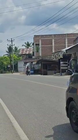 An ambulance with red flashing lights is driving down a road. The ambulance has text on the back that reads "RS ZAHRA LHOKSUKON". A white SUV is also driving on the road, behind the ambulance. The road is asphalt and there are trees and buildings on either side. The sky is cloudy.