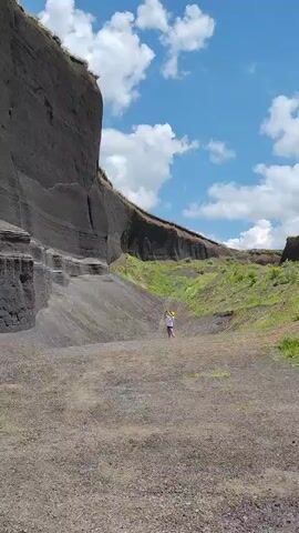 A person with light-colored clothing and yellow object in hand stands in a rocky, mountainous area. The person is walking in the distance toward the right side of the frame. The terrain is composed of gray and brown dirt and rocks with green foliage on top of a cliff in the background. The sky is blue with white clouds.