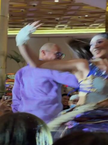 A man wearing a lavender shirt and glasses is on stage with a woman in an elaborate, sparkly blue and gold costume. The woman is performing a dance, and the man appears to be interacting with her, possibly also dancing or gesturing. The background suggests an indoor event or performance space, with a patterned ceiling and decorative columns. There are audience members visible in the foreground and background. The man is bald and smiling. The woman has her hair pulled back in a high ponytail and is looking towards the man. The image captures a moment of lively interaction between the two performers.
