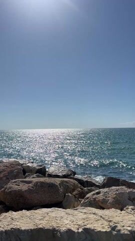 The video presents a serene, sun-drenched seascape. The scene is dominated by a vast expanse of calm, deep blue-green ocean under a clear, bright blue sky. The sun is visible in the upper left corner, casting a brilliant, shimmering reflection across the water, creating a sparkling path extending towards the horizon.

In the foreground, a collection of large, irregularly shaped rocks forms a barrier or shoreline. These rocks vary in color, with some appearing light beige or off-white and others a darker grey or brown. The water gently laps against these rocks, indicating a calm sea.

The overall impression is one of peacefulness and natural beauty on a clear, sunny day, likely during midday or early afternoon given the high position of the sun. There is no visible activity or people. The only sound present in the video is a soft, rhythmic lapping of ocean waves against the rocks, suggesting a gentle breeze and calm conditions. A distinct "ding" sound is heard at the very beginning of the video, which seems to be an external alert or notification sound rather than part of the natural environment.