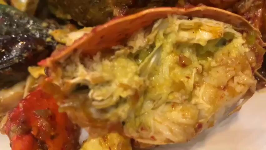 A close-up shot captures a generous platter of mixed seafood, served in a rich, reddish-orange sauce. The dish is a colorful assortment of crustaceans and mollusks, including bright red shrimp, segmented crab pieces with exposed white meat, and dark green mussels. Rings of squid are visible, intertwined with the other seafood. Corn on the cob, cut into thick rounds, is scattered throughout the platter, adding a yellow contrast to the predominantly red and green hues. The seafood appears glistening and saucy, suggesting it has been recently cooked and seasoned. A piece of crab shell, split open, reveals a pale green and yellow interior. The platter is white and round, resting on a wooden surface that might be a table. The lighting is warm and slightly diffused, creating a cozy atmosphere, possibly indicating an indoor setting, perhaps a restaurant. The overall impression is of a hearty and flavorful meal, likely a popular seafood boil or a similar communal dish enjoyed in a casual dining environment. There is no visible text in the media file.