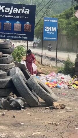 The video captures a roadside scene in Dhunibesi, Nepal, during the daytime under clear weather. In the foreground, a large pile of old, dirty tires is visible on a dirt patch, with a smaller stack of tires further right. To the right of the main tire pile, a substantial amount of colorful plastic garbage is strewn on the ground. A woman in a red and black checkered top is standing amidst the tires and garbage, appearing to sort through items or clean.

In the background, a paved road stretches, with vehicles, including motorcycles, occasionally passing by. Lush green hills or mountains rise in the distance. Two prominent billboards are visible. The one on the left advertises "Kajaria YOUR WORLDCLASS LUXURY," displaying images of decorative tiles and stating, "TRANSFORM YOUR SPACE INTO WORK OF ART." Below this in Nepali, it reads "बिस्तारै चलाऔं... सुन्दर घरले तपाईलाई कुरुदै।" which translates to "Let's drive slowly... A beautiful home awaits you." The billboard on the right indicates "MAW VRIDDHI 2KM AHEAD SPEED CHARGING." Power lines crisscross above the scene.