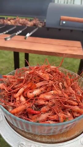 The video begins with a close-up of a glass bowl overflowing with cooked shrimp. The shrimp are bright orange and appear to be whole. In the background, a grill is visible with meat skewers cooking over charcoal. The grill is a dark color with a wooden table attached to its side. The table has a wooden top and metal legs. The scene is outdoors, with green grass visible in the foreground and background. The camera then pans to the right, focusing on the meat skewers on the grill. The meat is brown and appears to be well-cooked. The skewers are made of metal and have decorative handles. The video ends with a close-up of the meat skewers, showing the details of the cooked meat.