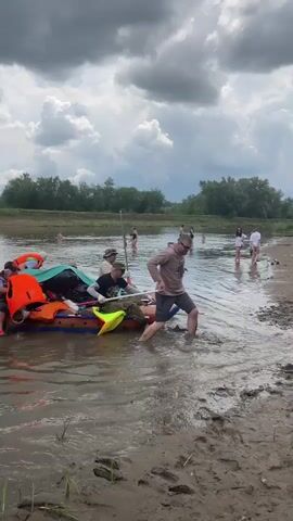 The video shows a group of people on a muddy riverbank preparing to launch an inflatable raft. Several individuals are in the water, some helping to push the raft, while others are already seated on it. The raft is loaded with gear, including life jackets. The sky is overcast with dark clouds, suggesting an impending storm. The surrounding environment is characterized by trees and vegetation along the river.