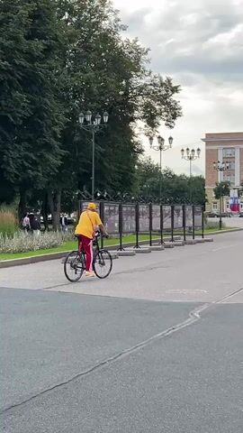 The media file shows a person riding a bicycle on a paved road in an outdoor urban setting. The individual is wearing a bright yellow t-shirt, red pants with white stripes, and a yellow cap. They are also carrying a red bag across their chest. The bicycle appears to be a black hybrid or commuter bike.

In the background, there is a large, ornate building with multiple windows and a light-colored facade, suggesting a public or official structure. Russian flags are visible hanging from the building. Several streetlights and trees line the area, indicating a park or boulevard. A row of display boards is also present along the side of the road. Other people and vehicles are visible in the distance, implying a moderately busy public space. The sky is overcast, suggesting cloudy weather. The overall scene appears to be set during daylight hours. The location is identified as Veliky Novgorod, Russia. There is no visible text in the media file.