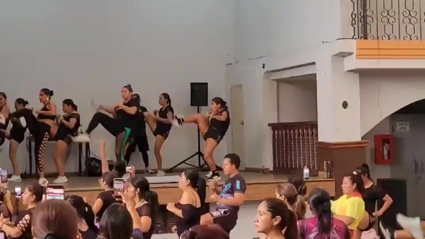 The video shows a group of women performing a choreographed dance routine on a raised wooden stage. The dancers are all wearing black athletic wear. There is an audience of people, mostly women, watching from below, many of whom are holding up their phones to record the performance. In the background, there are speakers and a laptop, suggesting the music is being played live or recorded. The environment appears to be an indoor venue, possibly a community hall or a gymnasium.
