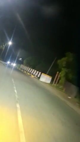 The video shows a nighttime view from a moving vehicle on a road. Headlights of approaching cars are visible in the distance, creating bright streaks of light. Streetlights illuminate the roadside, casting a warm glow on the asphalt. A concrete barrier with white and red reflective markers runs along the right side of the road, separating it from the dark surroundings, which appear to be lined with trees. The road itself is marked with a yellow line. The overall impression is of a journey through a dimly lit, semi-rural environment.