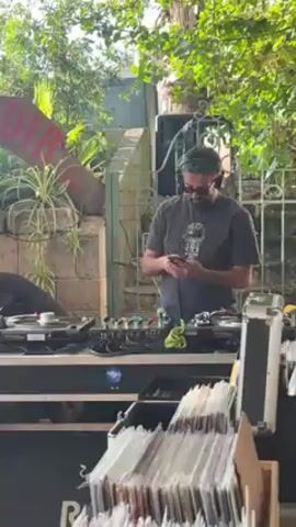 The video shows a man using his smartphone while standing behind DJ equipment. He is wearing sunglasses and headphones. There are two record players, a mixer, and crates of records set up on a table. Another person is behind one of the record players, wearing black. The setup is outdoors, surrounded by trees and greenery, in what appears to be a garden or backyard setting. A sign that says "DIRT" is slightly visible in the background.