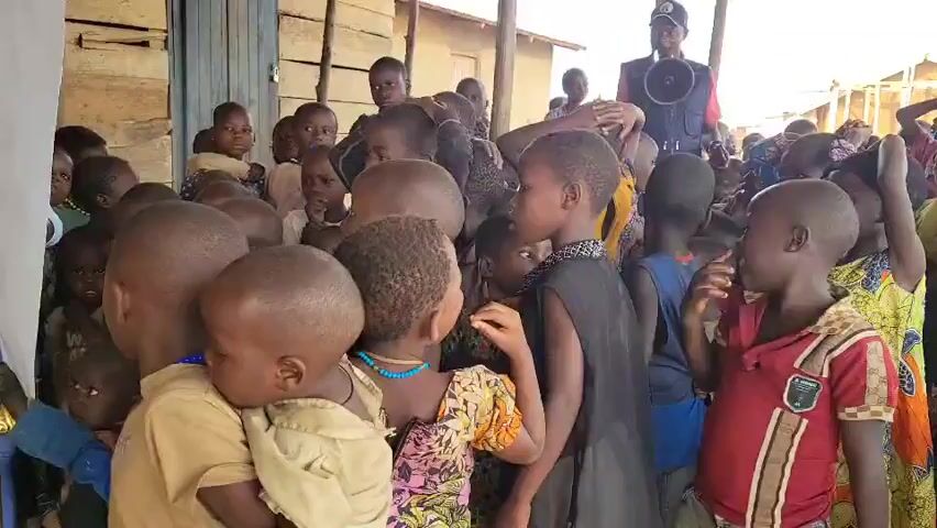 A group of young children are gathered in what appears to be an outdoor setting, possibly a temporary shelter or settlement. The children are of varying ages, with many appearing to be toddlers and pre-schoolers. They are clustered together, some looking towards the camera, others interacting with each other or looking off-frame. The background consists of simple wooden structures, suggesting a rural or underdeveloped area. In the background, a person is visible holding a megaphone, and another person appears to be taking a photo. The children are dressed in simple clothing. Some of the children are wearing necklaces. The overall impression is of a community gathering or an event for children.
