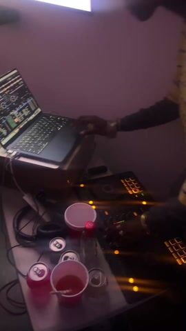 The scene captures a person, likely a DJ, working with electronic equipment. The individual is partially visible, wearing a yellow patterned shirt with black sleeves and adorned with jewelry on their hands and wrist. They are positioned in front of a DJ controller with illuminated buttons. A laptop sits to their left, displaying a screen with waveform visualizations, suggesting music production or mixing software. The scene is slightly blurred, but visible objects on the table also include headphones, several opened and unopened cans of a red beverage, and red plastic cups, one containing a reddish liquid and a straw. The background is nondescript, suggesting an indoor, possibly dimly lit environment.