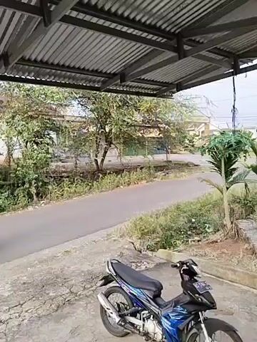 The video begins showing a blue and black motorcycle parked on a concrete surface. In the background, a paved road curves to the left, bordered by greenery and trees. Houses with tiled roofs are visible behind the vegetation. A corrugated metal roof supported by metal beams extends over the foreground. At the 00:01 mark, a motorcycle carrying two individuals approaches from the distance on the paved road. The driver is wearing a light-colored shirt, and the passenger appears to be wearing a dark top and light-colored pants. They are riding away from the viewer. At the 00:02 mark, the motorcycle with two riders continues to move away. At the 00:03 mark, the motorcycle with two riders is still visible on the road, heading away from the camera. At the 00:04 mark, the road is empty again, and the motorcycle in the foreground is still parked. The scene is outdoors during daylight.