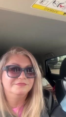 A young blonde woman in sunglasses is in the passenger seat of a car, filming herself. She has long blonde hair, is wearing sunglasses with dark frames and pink lenses, and a black shirt over a pink top. Behind her, in the back seats, are two other blonde women, also wearing sunglasses. The woman in the middle back seat is wearing a white t-shirt and has a beige handbag on her lap. The woman in the right back seat is wearing a light blue collared shirt. All three women have blonde hair. The car's ceiling is visible, with a warning label about airbags and children in the back seat. There are reflections of the outside environment visible in the sunglasses of the women.