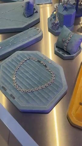 The camera scans across a display of jewelry on a silver-colored surface. Several velvet display pads hold various necklaces, bracelets, and earrings. Some of the pads are a light blue, while others are a tan color with a quilted diamond pattern. Many of the jewelry pieces feature colorful gemstones. Price tags are visible next to some of the items. The lighting in the scene is bright and somewhat bluish.