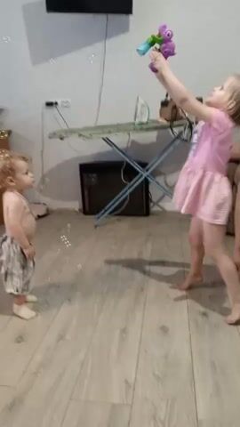 A young boy with curly hair and wearing patterned shorts stands facing a young girl in a pink dress. The girl is holding a toy that is producing bubbles, and she is looking at the boy. The boy is looking up at the bubbles and pointing. The scene appears to be indoors in a room with wooden floors. There is an ironing board in the background with items on it.