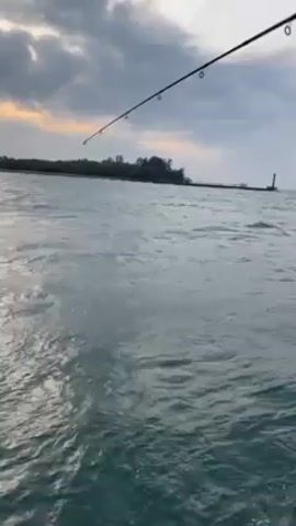 The video shows a view from a boat with a fishing rod pointing out over the water. The water is choppy, and there is an island with a lighthouse visible in the distance under an overcast sky. The boat appears to be moving through the water, creating waves.
