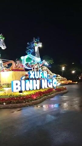 At night, the video shows a brightly lit display with two horse sculptures made of a wireframe construction and illuminated with lights. The horses appear to be hitched to a carriage-like structure on the left, which is also brightly lit. In front of the horses, there is a raised platform spelling out "XUAN BINH NGO" in large, blue illuminated letters. The letters are surrounded by colorful flower beds. The ground appears wet, suggesting it might have rained recently. Background elements include trees and other lights. The overall scene suggests a celebratory or festive display.