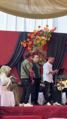 The video shows a band performing at a wedding reception. The band consists of at least six members. The lead singer is a man in a white striped shirt and dark pants. Next to him is a man in a dark vest, glasses, and a green shirt, also singing. To the left is a man with a guitar, wearing a green shirt, black pants, and a baseball cap. Another band member is playing keyboard and wearing sunglasses. To the right of the lead singer is a man playing guitar. There is also a drummer. The stage is decorated with an array of flowers. A seated woman wearing a green hijab and embroidered dress sits to the side of the stage. The background is draped with black and red fabric. The audience is visible only partially at the bottom of the frame. The setting indicates a festive, celebratory occasion.