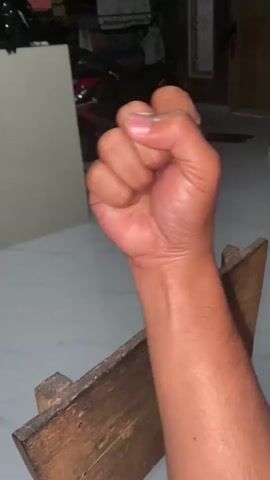 The video shows a close-up of a person's arm and hand. The hand starts in a fist, then opens into a palm, then forms a thumb gesture, and finally returns to a fist. The arm is hairy and tanned. The person is sitting and the arm is resting on a wooden object. The background is out of focus but appears to be an indoor setting with furniture and possibly a motorcycle. The lighting is dim.