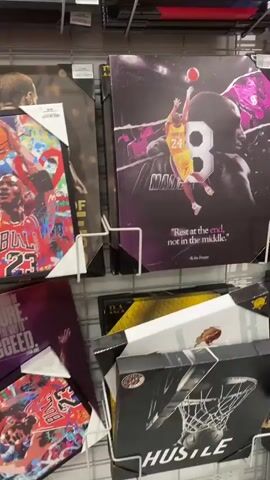 The video shows a wall display of framed artwork, likely in a retail store. The artwork features prominent basketball players. In the foreground, a framed print depicts Kobe Bryant in a yellow jersey with the number 24, mid-air reaching for a basketball. Behind him, a large number 8 is visible, along with a quote attributed to Kobe Bryant: "Rest at the end, not in the middle." To the right of this print, a vibrant, abstract-style print shows Michael Jordan in a red jersey with the number 23, also holding a basketball. Other framed prints are partially visible above and below these central pieces, including a colorful, abstract piece at the top and another print featuring a basketball player in the lower portion of the frame. The overall impression is a collection of sports-themed art.