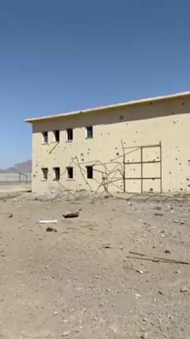 The video shows a damaged building, pockmarked with bullet holes and with the upper part of the roof significantly damaged. A burned-out car sits in the foreground amid debris and a man in a brown robe walks across the landscape in front of the building. The sky is clear and blue. The landscape is arid and appears to be in a state of disrepair.