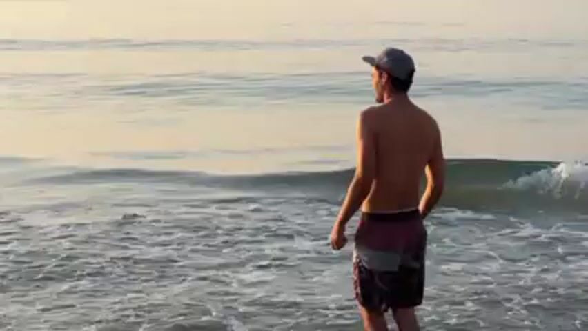 A shirtless man wearing a baseball cap and patterned shorts stands at the edge of the water at a beach. He tosses a disc into the water. The camera follows the disc's trajectory as it skips across the surface of the water. The backdrop is a calm ocean during sunrise or sunset. Other people and distant boats are visible on the horizon.