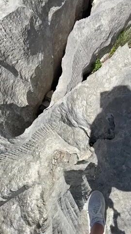 This media file shows a close-up, high-angle view of a rugged, grey, rocky landscape. The rock formations are uneven with deep crevices and sharp edges. Some areas of the rock have a rough, textured surface, appearing almost carved or scored, while others are smoother with patches of white discoloration. Small tufts of green grass and vegetation are visible growing in some of the cracks and crevices, suggesting an outdoor environment.

In the lower right of the frame, a person's right foot wearing a white sneaker is visible, indicating the perspective is from someone standing on the rocks. The shadow of a person holding a phone or camera is cast prominently on the rocks, suggesting the photo was taken by someone documenting their experience. The lighting suggests it is daytime, likely sunny, given the clear shadows.

The overall impression is of a natural, somewhat harsh, geological environment. The textures of the rock and the presence of vegetation hint at a remote or mountainous area. The image does not contain any visible text. The media appears to capture a moment of exploration or hiking in this distinct natural setting.