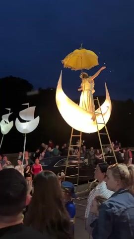 A nighttime outdoor event is taking place, with a crowd of people gathered. A performer stands on a crescent moon structure, holding a yellow umbrella. The moon structure is lit and has a ladder leading up to it. In the background, there are other illuminated floats or decorations, including two ship-like structures on the left and a pink and yellow domed structure on the right. The crowd is looking towards the performer and the floats, with some people holding up phones to take pictures or videos. The atmosphere appears festive.