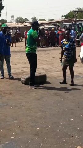 The video depicts three individuals in an outdoor setting with a gathering of people in the background. The environment is somewhat rough, resembling a dusty or unpaved area with buildings and vehicles visible. A man wearing a green shirt and a cap stands on a large tire, speaking into a microphone. Another man stands to his left, holding a camera and filming. A woman stands on the right, holding papers or a small book. The gathering of people in the background seems to be watching or observing the scene.