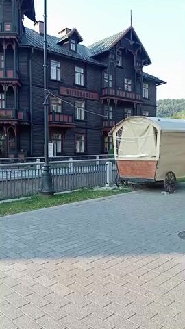 The video pans across the exterior of several large, multi-story wooden buildings, likely hotels or inns, with ornate architecture and balconies. In the foreground, a paved walkway is visible, bordered by a low fence and grass. A single lamppost stands near the fence. To the right, a small, covered wagon or trailer is parked. The buildings are dark wood with red trim and light-colored window frames. Sunlight illuminates parts of the scene, suggesting it is daytime. No specific actions or interactions between people are clearly discernible beyond brief glimpses of individuals near the building entrances. The overall impression is of a historical or resort-like setting.