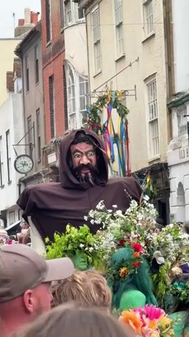 A low-angle shot captures a procession in a densely built urban environment. In the center is a large, imposing figure of a man with dark skin, a beard, and a hooded brown robe. The figure appears to be part of a float or display, surrounded by greenery and white flowers. Below the figure, several people are visible with their faces painted green and wearing floral headdresses. A crowd of onlookers, partially visible in the foreground, watch the procession. The buildings lining the street are varied in architectural style, with brick and painted facades, multiple windows, and a clock mounted on one building. A sign reading "Electric Palace Cinema" is also visible. The overall scene suggests a festive event, possibly a parade or celebration.