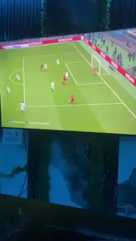 A television is showing a soccer video game. The game depicts a match between two teams, one in white and one in red. The red team is attacking the goal. The white team's goalie dives to block a shot. The setting appears to be someone's living room with a wall mural of mountains in the background.