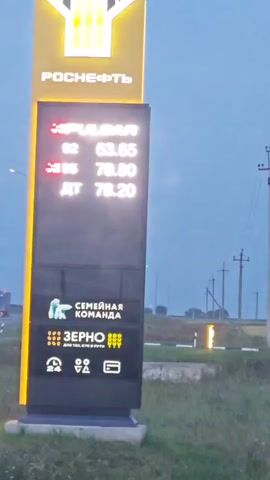 A digital price board for a Rosneft gas station is shown. The board displays fuel prices including Pulsar 92 at 63.65, 95 at 70.80, and Diesel at 78.20. Below the fuel prices, there are logos and text indicating "Family Team" and "Grain". The bottom section of the board shows various icons, including a clock with 24, a car with a person, and a receipt. The sky is overcast. In the background, a road with a truck can be seen.