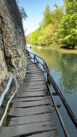 The video shows a slightly elevated wooden plank walkway on the side of a rocky cliff, bordering a river. The walkway has a wooden railing. A group of people are walking on the walkway. In the river, some people are floating in inflatable rafts. The river is surrounded by green trees on the opposite bank, and the sky is clear and blue. The sun is shining.