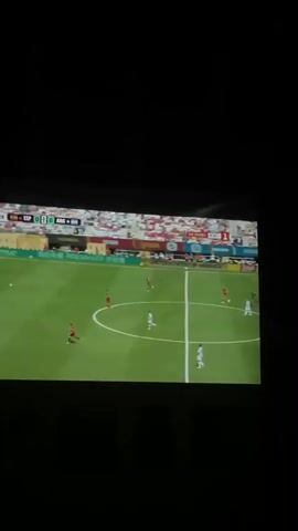 The video shows a projection of a soccer game on a screen. The game is being played on a green field with white lines. There are several players on the field, wearing red and white uniforms. In the background, there are spectators in the stands. There is text overlaid on the bottom of the screen in a foreign language, which appears to be Bulgarian. The score on the screen reads 0-0. The time displayed is 29:31. The camera is positioned in front of the screen and is angled slightly upwards. The scene is dimly lit, with the screen being the primary light source.