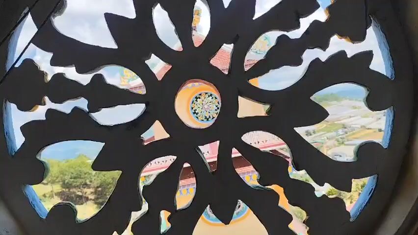The video displays a view through a dark, intricate, sunburst-shaped cutout in what appears to be a wall or screen. The cutout reveals a landscape beyond, featuring a blue sky with white clouds, green hills, and a distant town or village with numerous greenhouses. Architectural elements of a building, possibly a temple or pagoda with colorful tiled roofs and ornate details, are visible through some of the openings in the cutout. The central part of the cutout reveals a circular, decorative element with colorful geometric patterns. The camera appears to be stationary, offering a static perspective of this scene. There are no visible people or discernible actions taking place.