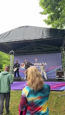 The video shows an outdoor stage with a band performing, as viewed from a few spectators standing in the grass. A female singer in a black jacket is at the center of the stage, accompanied by a guitarist to her left and a dancer to her right. The stage has a black roof and a purple front with white text and an emblem. Several people are in the grass near the stage, some holding papers. A wooded area is in the background. The sky is overcast.