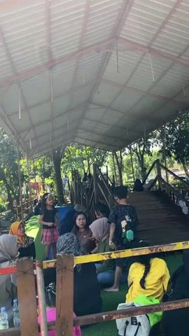 This image depicts a group of people gathered under a covered outdoor structure. The subjects include children and adults, some of whom are wearing hijabs. They appear to be seated on a green surface that resembles artificial grass, with some on a wooden platform. In the background, there's a wooden walkway or bridge leading up to more elevated structures, suggesting a park or recreational area. Trees and greenery are visible beyond the covered area, indicating an outdoor setting.

The activity seems to be a casual gathering or break, possibly during an outing or event. Some individuals are looking at each other, engaged in conversation. The lighting suggests it's daytime, with sunlight filtering through the trees and the open sides of the structure. The weather appears to be pleasant and sunny.

Notable details include a young girl in the foreground wearing a red and plaid skirt and a flower crown, and a boy in the background wearing a t-shirt with a graphic. There are plastic bottles and bags visible, suggesting personal belongings. The overall mood seems relaxed and informal. There is no visible text in the image.