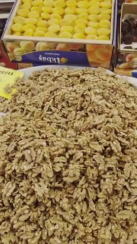 The video pans over a market stall filled with dried fruits and nuts. In the foreground, a large pile of shelled walnuts is displayed in a white bowl. Behind the walnuts, there are boxes filled with dried apricots, some appearing bright yellow and others a darker brown. Further back, a box of dark brown dates is visible, along with containers of other dried goods. Some packaged items, resembling walnut strings covered in a dark coating, are also in the foreground. The lighting suggests an indoor or covered market setting. The camera movement is smooth, showcasing the variety of products.