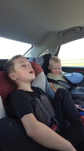 Two young boys are sitting in the back of a car, presumably on a journey. The boy in the foreground, wearing a black t-shirt, appears to be yawning or singing. He is seated in a car seat or booster seat with a grey harness. Behind him and to the right, another boy with blond hair, wearing a light green t-shirt, is also in a car seat with a blue headrest. He is smiling and looking towards the camera, and briefly raises his right hand as if to wave. The interior of the car is visible, with the ceiling and part of the car door. Outside the windows, a landscape with green fields and a bright sky can be seen. The lighting suggests it is daytime.