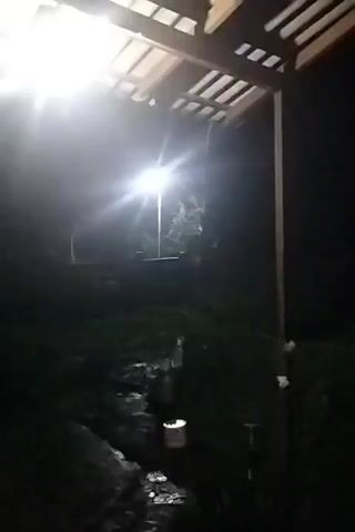 The video begins with a view looking out from under a covered structure. A bright light illuminates the area, casting a glare. The ground appears wet and reflects the light, suggesting it may have rained or is raining. A path or walkway is visible, also wet. Further in the background, trees and vegetation are dimly visible in the darkness. A lamp post with a light is also seen. Towards the right side of the frame, a scooter is parked near a pole. The scene is dark, with the artificial light being the primary source of illumination. The video is brief and appears to be handheld, with some camera movement.