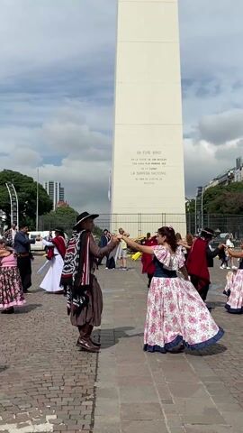 The video captures a group of people dancing in front of the Obelisco de Buenos Aires, a prominent landmark in Argentina. The dancers, both men and women, are dressed in traditional Argentine clothing. The men wear hats, ponchos, and boots, while the women are in long, floral dresses. They move in a synchronized manner, reflecting a cultural dance. The obelisk stands tall in the background, with inscriptions visible on its surface. The environment includes a paved square with other people walking around, and some vehicles can be seen nearby. The sky is partly cloudy, suggesting daylight.