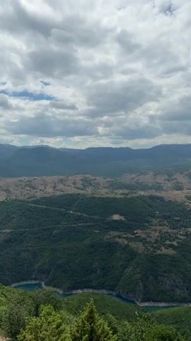 A drone flies over a vast mountainous landscape with a winding blue river flowing through it. The sky is partly cloudy. The terrain is covered with lush green forests and some sparsely vegetated hills. The drone captures a panoramic view of the valley, showcasing the grandeur of the natural environment.