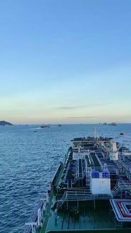The video shows a view from the deck of a large cargo ship at sea, likely a tanker. The ship is moving through calm, blue water under a clear, pale blue sky with a faint horizon line suggesting either dawn or dusk. The deck of the ship is visible in the foreground, featuring various pipes, railings, and equipment. Some of these pipes are colored red and white. There are also several large cylindrical tanks on the deck.

In the middle ground and background, the sea is dotted with other vessels, including several cargo ships and smaller boats. One ship in the distance appears to be moving at a faster speed, leaving a white wake. Land is visible on the far left of the frame, appearing as a low-lying island or coastline.

The time of day appears to be early morning or late evening, given the soft lighting and the color of the sky. The weather seems pleasant with minimal clouds. The overall mood is serene and expansive, emphasizing the vastness of the sea and the scale of maritime transport.

Visible text on the ship includes a sign that reads "NO SMOKING SAFETY FIRST." The text is white on a black background.

The context provided indicates the location is Penajam, Indonesia, suggesting the ship is likely in Indonesian waters. The presence of numerous ships could indicate a busy shipping lane or a port area.