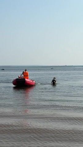 A red inflatable boat with two people is in the water. One person, wearing an orange life jacket, is seated. The other person, wearing a white shirt, is also seated. A third person, wearing a dark swimsuit, is standing in the water to the right of the boat, splashing their arms. The water is calm, with small ripples. The sky is clear and blue. In the distance, there are faint shapes of other people or objects in the water.