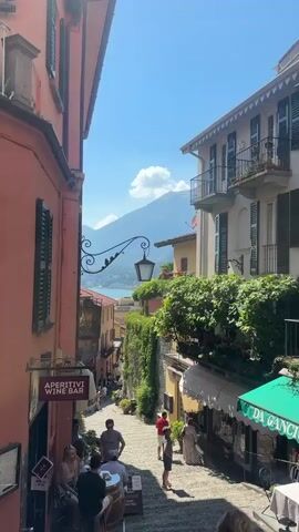 This media file captures a picturesque street scene in San Giovanni, Italy. The image is dominated by a steep, cobblestone alleyway lined with buildings on either side. Verdant greenery cascades from balconies and walls, adding a vibrant, natural element to the architecture. In the distance, a tranquil lake is visible, framed by majestic, mist-shrouded mountains under a bright blue sky dotted with fluffy white clouds.

Several people are scattered throughout the alley, engaged in various activities. Some are seated at outdoor tables, enjoying refreshments, while others are strolling up or down the incline. The atmosphere appears relaxed and leisurely, suggesting a pleasant day for sightseeing or dining.

Notable details include a vintage-style street lamp suspended from an ornate iron bracket, adorned with decorative bird silhouettes. Several buildings feature traditional shutters and balconies, characteristic of Italian architecture. A sign above an establishment reads "APERITIVI WINE BAR," indicating a place for drinks and socializing. Another awning displays "DA GANGI ITALIAN STYLE," suggesting a restaurant.

The time of day is clearly daytime, with strong sunlight casting shadows and illuminating the scene. The weather is fair and warm, conducive to outdoor activities. The combination of charming architecture, lush vegetation, and the stunning natural backdrop of the lake and mountains creates a sense of idyllic beauty and timeless charm. The overall emotion conveyed is one of peaceful enjoyment and the simple pleasures of a beautiful Italian locale.