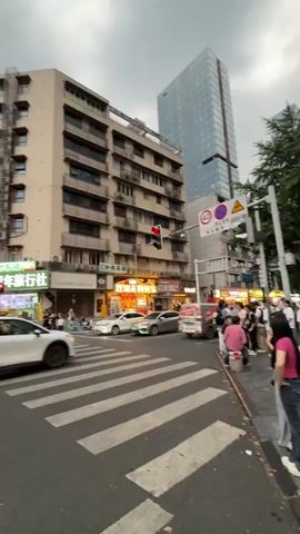The video shows a busy city intersection in a medium shot. The sky is overcast and grey. In the foreground, cars and motorbikes are visible, with several people riding motorbikes. A white crossover SUV drives past in the foreground. In the background, there are multi-story buildings, some with advertisements and signs, and a tall modern skyscraper. Traffic lights are visible at the intersection. The street has zebra crossings.