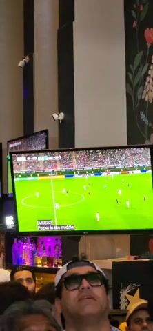 A group of people are watching a soccer game on multiple screens in a dimly lit establishment. The screens show a green soccer field with players. The people in the foreground are looking up at the screens, some with their mouths open in excitement or surprise. The environment appears to be a bar or restaurant with bottles visible behind the counter. The atmosphere is lively and engaged with the game.