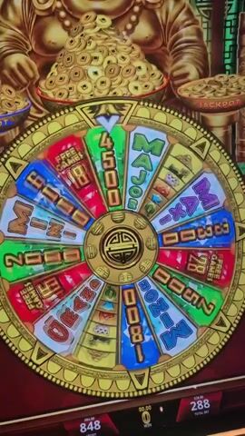 The video shows a close up of a slot machine with a bonus wheel, adorned with a smiling golden Buddha statue at the top. The wheel is multicolored and sectioned, featuring prizes like "MINI," "MAXI," "GRAND," "2500," "3300" and "Free Games." The wheel is actively spinning. Below the wheel, the machine displays digital readouts showing the current credit balance, the total bet amount, and any winnings. The environment is what looks to be a casino, as evidenced by the slot machine.