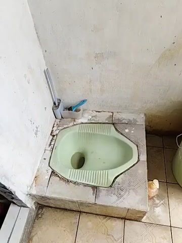 A squat toilet is in a tiled bathroom. The toilet is green and has a light green rim. There is a toilet brush and a blue toothbrush holder next to the toilet. A large green bucket with a blue ladle is to the right of the toilet. A pipe with a red valve extends from the wall and is connected to a hose that goes into a white bucket. The floor is tiled with a pattern of white and light gray squares. The walls are painted a light color and appear to be stained in places. There is a white door to the right of the scene.